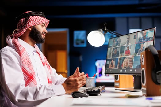 Confident Arab businessman in traditional clothing engaged in online video conferencing at a desk. Muslim guy listens attentively to colleagues, using desktop pc for communication and research.