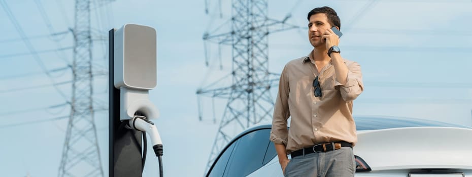 Man talking on the phone while recharge EV car battery at charging station connected to power grid tower electrical as electrical industry for eco friendly car utilization. Panorama Expedient