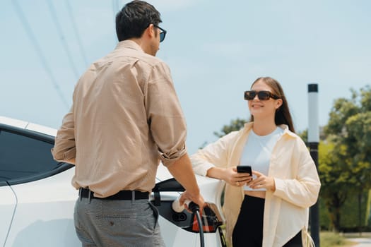 Young couple use smartphone to pay for electricity at public EV car charging station green city park. Modern environmental and sustainable urban lifestyle with EV vehicle. Expedient