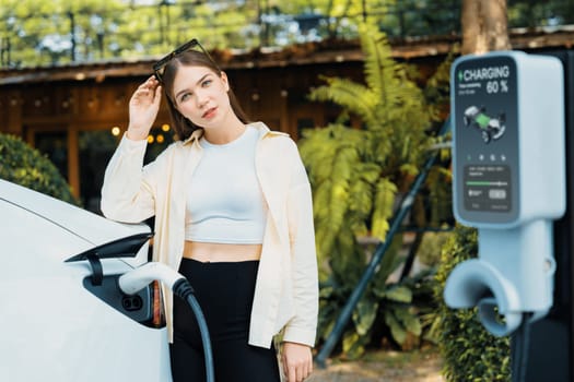 Young woman and sustainable urban commute with EV electric car recharging at outdoor cafe in springtime garden, green city sustainability and environmental friendly EV car. Expedient