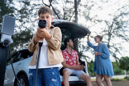 Little boy holding EV charger and point at camera with his family sitting on the trunk in background. Road trip travel with alternative energy charging station for eco-friendly car concept. Perpetual