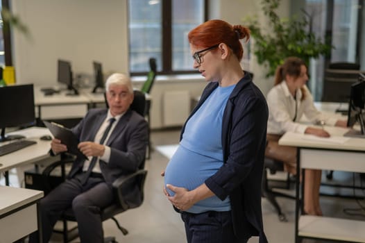 A pregnant woman suffers from pain and holds her stomach while standing in the middle of the office next to her colleagues