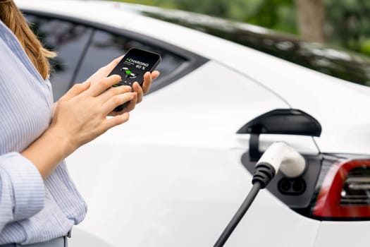 Young woman using smartphone online banking application to pay for electric car battery charging from EV charging station during vacation holiday road trip at national park or summer forest. Exalt