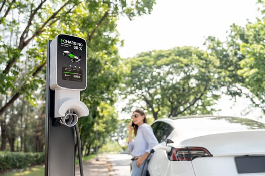 Young woman using smartphone online banking application to pay for electric car battery charging from EV charging station during vacation holiday road trip at national park or summer forest. Exalt