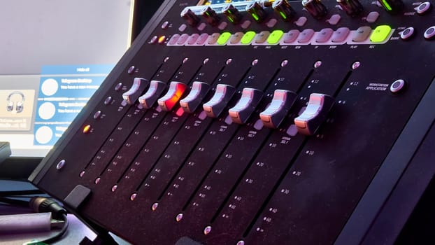 Low angle close up on a dark studio mixing sound board, with rows of small dials illuminated by a rainbow spot light, in the control booth at a concert