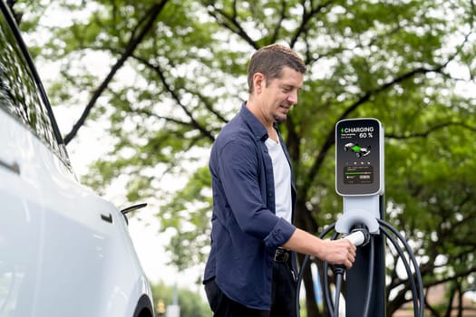 Man recharge EV electric vehicle's battery from EV charging station in outdoor green city park scenic. Eco friendly urban transport and commute with eco friendly EV car travel. Exalt