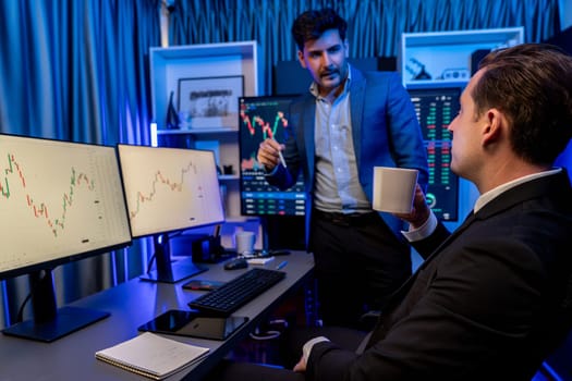 Two stock exchange traders discussing dynamic investment graph in currency rate on monitor at night. Businessman partners coffee meeting in high stock market in neon light at workplace. Sellable.