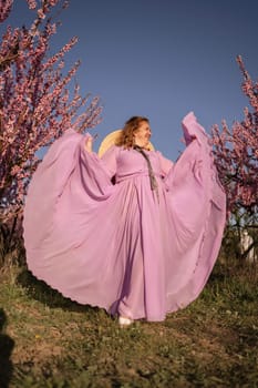 Woman blooming peach orchard. Against the backdrop of a picturesque peach orchard, a woman in a long pink dress and hat enjoys a peaceful walk in the park, surrounded by the beauty of nature