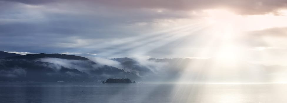 Sun rays, sea and clouds in blue sky with mountains, water and fog in evening. Lake, landscape and environment for holiday in summer, travel and destination for vacation or peace with scenery.