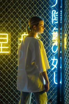 A beautiful blonde girl in a white oversized T-shirt and blue jeans posing against the background of neon hieroglyphs. High quality photo