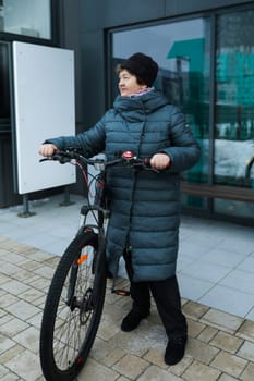 Mature European woman dressed in a winter coat rides a bicycle along the city streets.