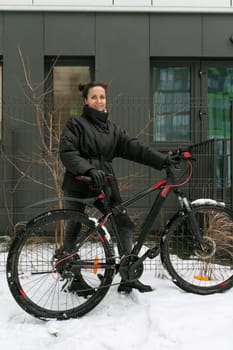 A pretty brunette woman stands with a sports bike in the yard of her house.