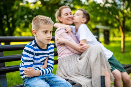 Happy mother is sitting with her sons on bench in park. One boy is offended and pouting.