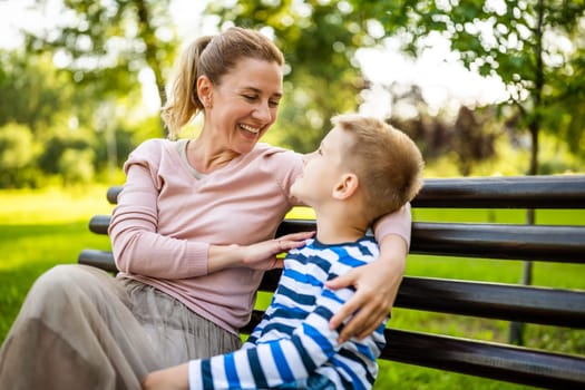 Happy mother is sitting with her son on bench in park. They are having fun together.