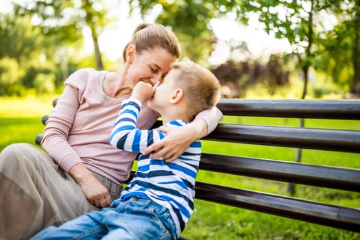 Happy mother is sitting with her son on bench in park. They are having fun together.