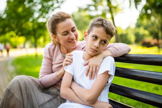 Happy mother is sitting with her son on bench in park. Boy is offended and pouting.