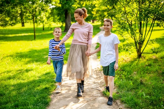 Happy mother is walking with her sons in park. They are holding hands and enjoying summer day.