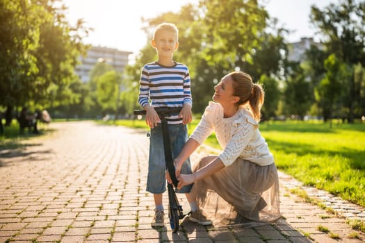 Mother having fun with her son in park on sunny day. Boy is ridding scooter.