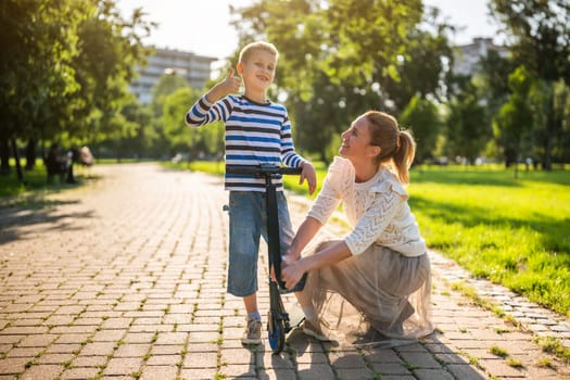 Mother having fun with her son in park on sunny day. Boy is ridding scooter.