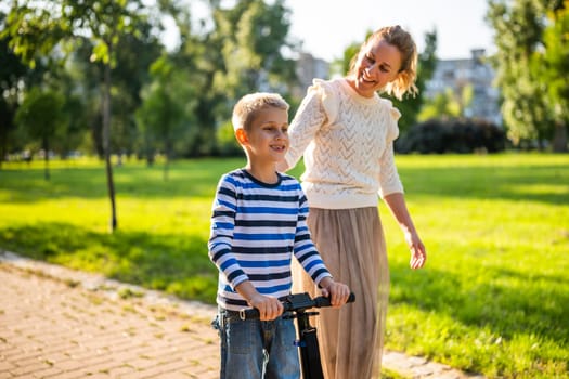 Mother having fun with her son in park on sunny day. Boy is ridding skateboard and his mother is pushing him.