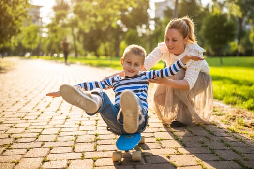 Mother having fun with her son in park on sunny day. Boy is ridding skateboard and his mother is pushing him.