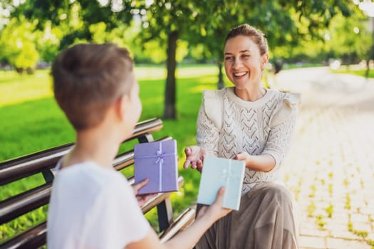 Happy mother is sitting with her son on bench in park. Boy is giving a present to his mother.