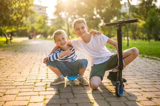 Two boys are having fun with skateboard and scooter in park. Playful children in park, happy childhood.