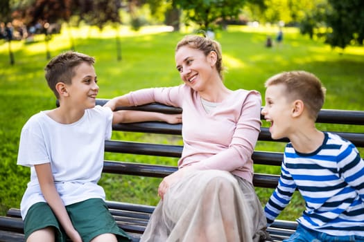 Happy mother is sitting with her sons on bench in park. They are talking and enjoying their time together.