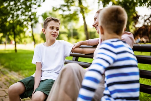 Happy mother is sitting with her sons on bench in park. They are talking and enjoying their time together.