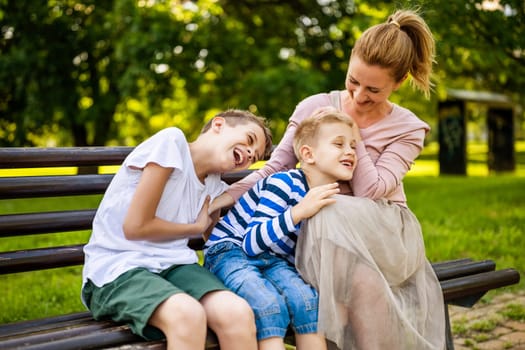 Happy mother is sitting with her sons on bench in park. They are talking and enjoying their time together.