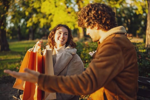 Couple sitting on bench in park. Woman is showing clothes to her man after shopping.