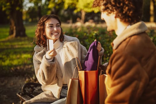 Couple sitting on bench in park. Woman is showing clothes to her man after shopping.