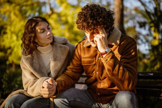 Young couple is sitting in park on sunny day. Man is sad and woman in consoling him.