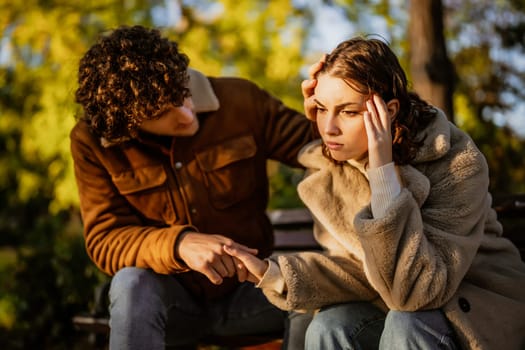 Young couple is sitting in park on sunny day. Woman is sad and man in consoling her.