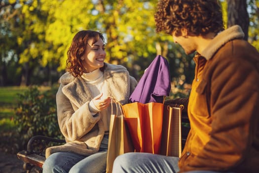 Couple sitting on bench in park. Woman is showing clothes to her man after shopping.