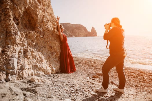 Side view a Young beautiful sensual woman in a red long dress posing on a rock high above the sea during sunrise. Girl on the nature on blue sky background. Fashion photo.