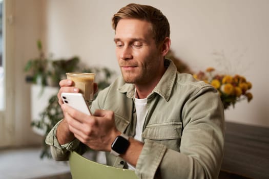 Lifestyle portrait of handsome young man, sitting in cafe, checking his phone and drinking coffee, smiling. Concept of leisure, going out