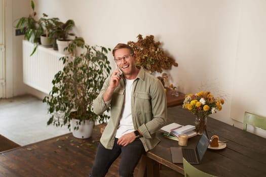 Portrait of handsome happy man in an office, guy sits in coworking space, looking at camera, smiling and laughing, talking on mobile phone, using smartphone, answer a call from client.
