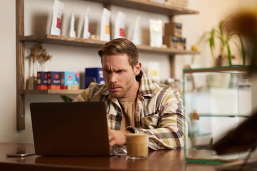 Portrait of businessman in cafe, looking serious at his laptop, working on project with concentrated face expression, has remote job, sitting in coffee shop.