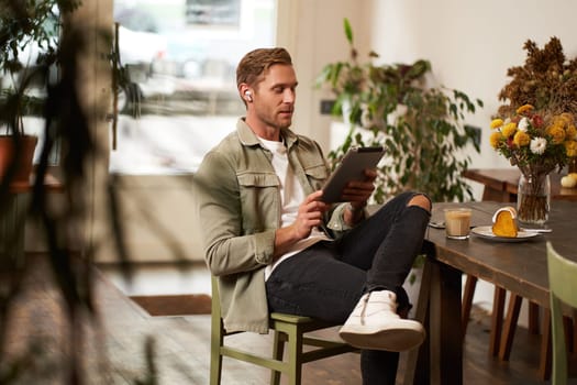 Lifestyle shot of handsome young man sitting in a cafe in front of the table, drinking coffee, wearing wireless headphones, listening to music or watching video in public space, using digital tablet.