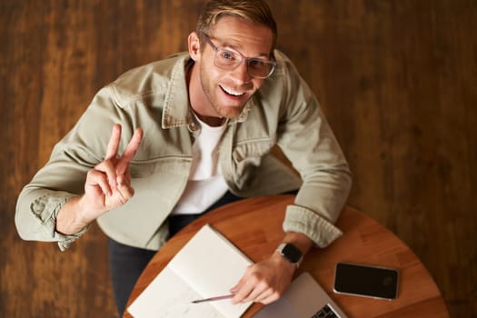 Upper angle shot of handsome smiling man in glasses, showing peace sign and smiling at camera, working in cafe, studying in front of laptop, reading notes in notebook.