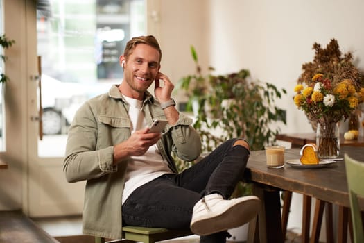 Lifestyle portrait of handsome stylish young man, sits in cafe, holds mobile phone, puts on wireless earphones, looks at camera with happy smile.
