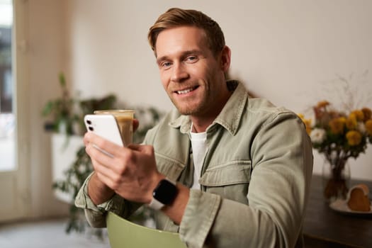 Lifestyle portrait of handsome young man, sitting in cafe, checking his phone and drinking coffee, smiling. Concept of leisure, going out