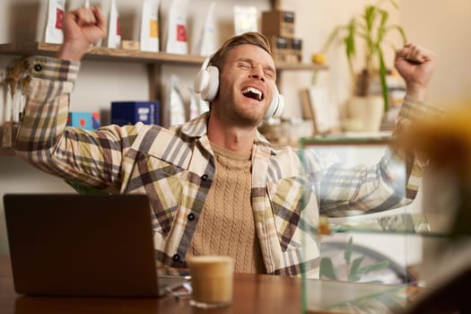 Portrait of excited, happy young man sitting behind the counter in cafe, working on laptop, drinking coffee, listening music in wireless headphones, singing high note.