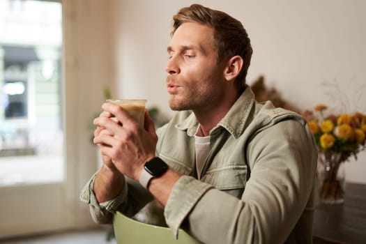 Portrait of young handsome bearded man in coffee shop, sitting on chair, sipping cappuccino, resting in cafe.