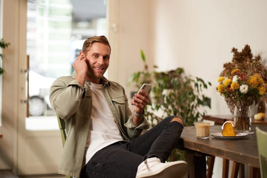 Happy handsome man in coffee shop, listening to music in wireless earphones, connects to cafe wifi to enjoy favourite song on his smartphone playlist, sitting and drinking cappuccino.