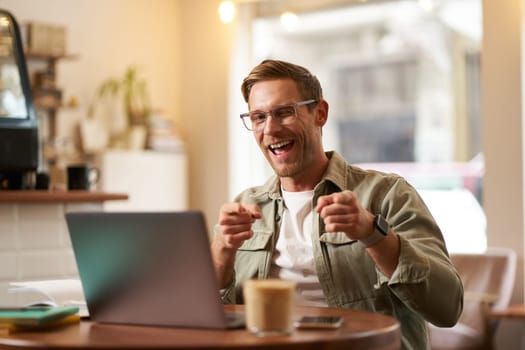 Happy young man in glasses, freelancer sitting in cafe with laptop, drinking coffee, pointing fingers at camera and smiling cheerfully.