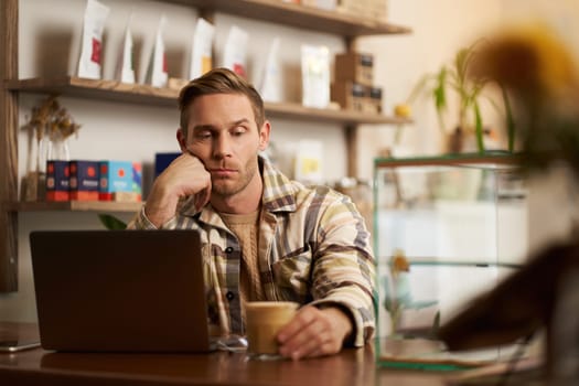 Portrait of man looking sad and stressed at his glass of coffee, working from cafe, sitting with laptop, lacks inspiration, doesnt feel inspired to finish the project.