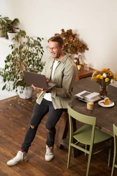 Vertical portrait of handsome happy man, manager or CEO of company, sitting on table and holding laptop, laughing, video chatting, has an online meeting with a team, standing in an office.