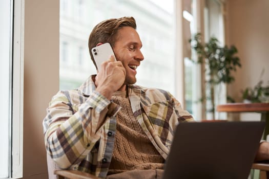 Image of happy cafe visitor, man with laptop, calling a friend over the phone. Businessman talking on telephone, working online from coffee shop, laughing and smiling.